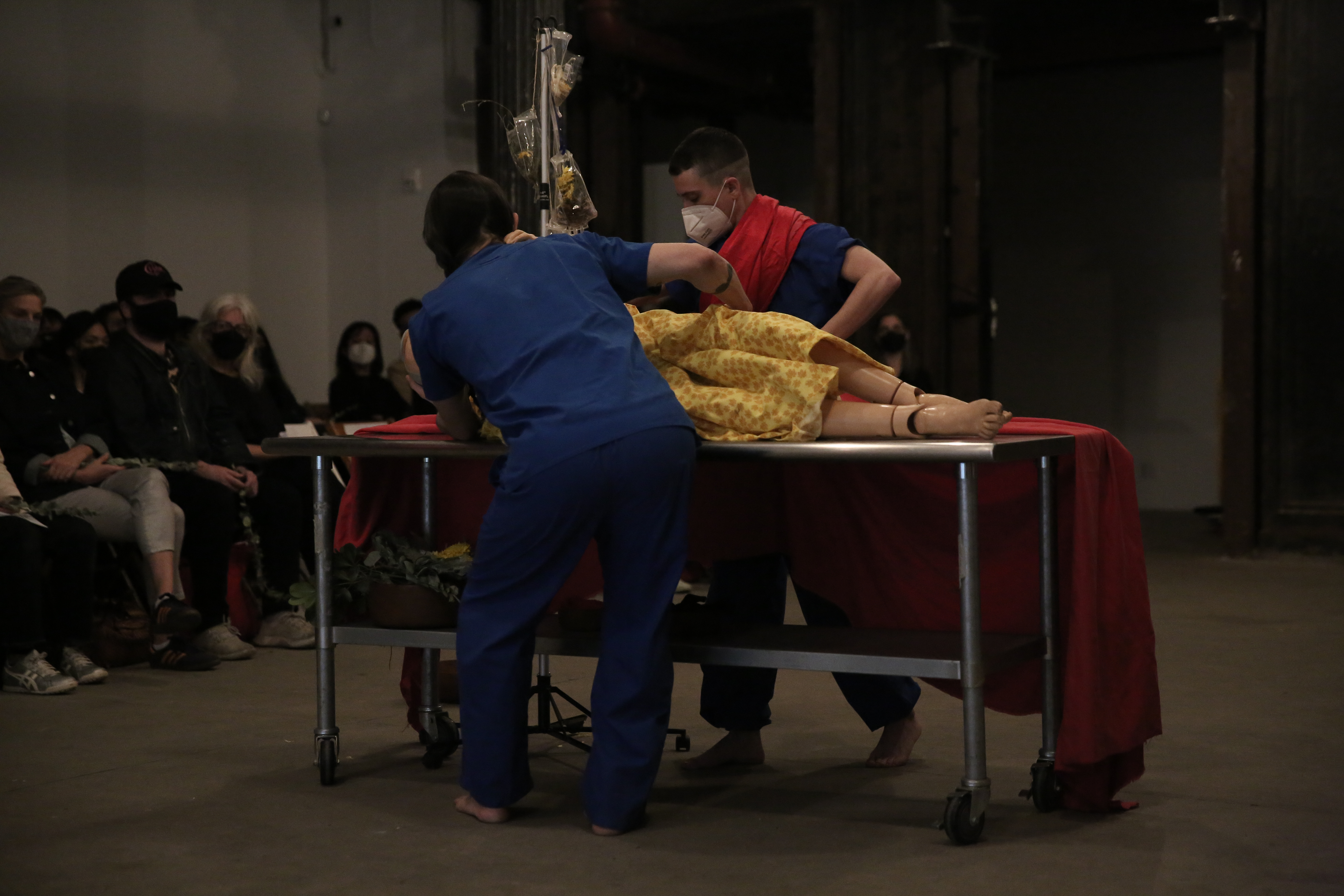 Two figures wearing medical scrubs move a table with wheels, on which a mannequin lies.