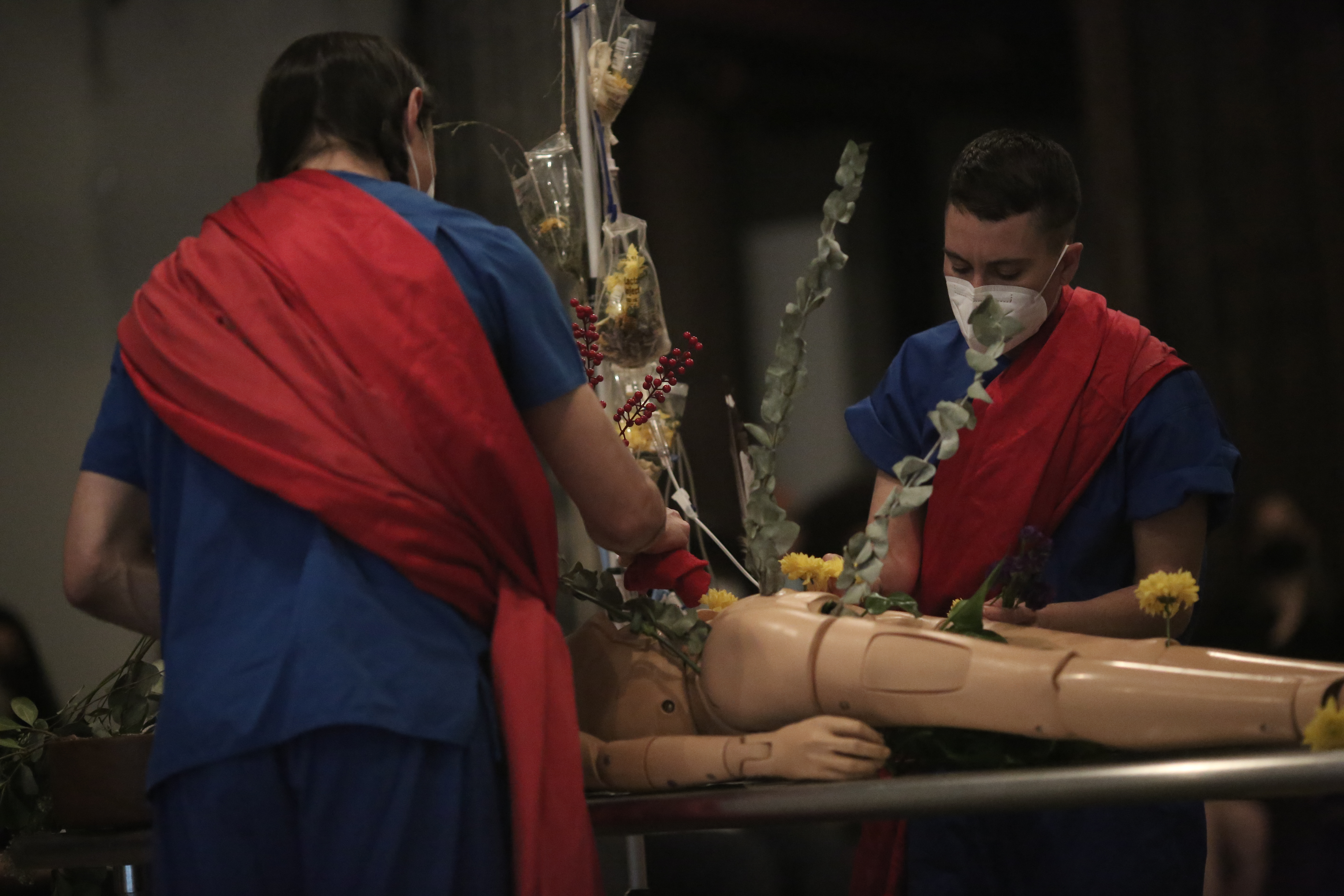 Two figures hold medical instruments over a table with a manneuqin and an IV.