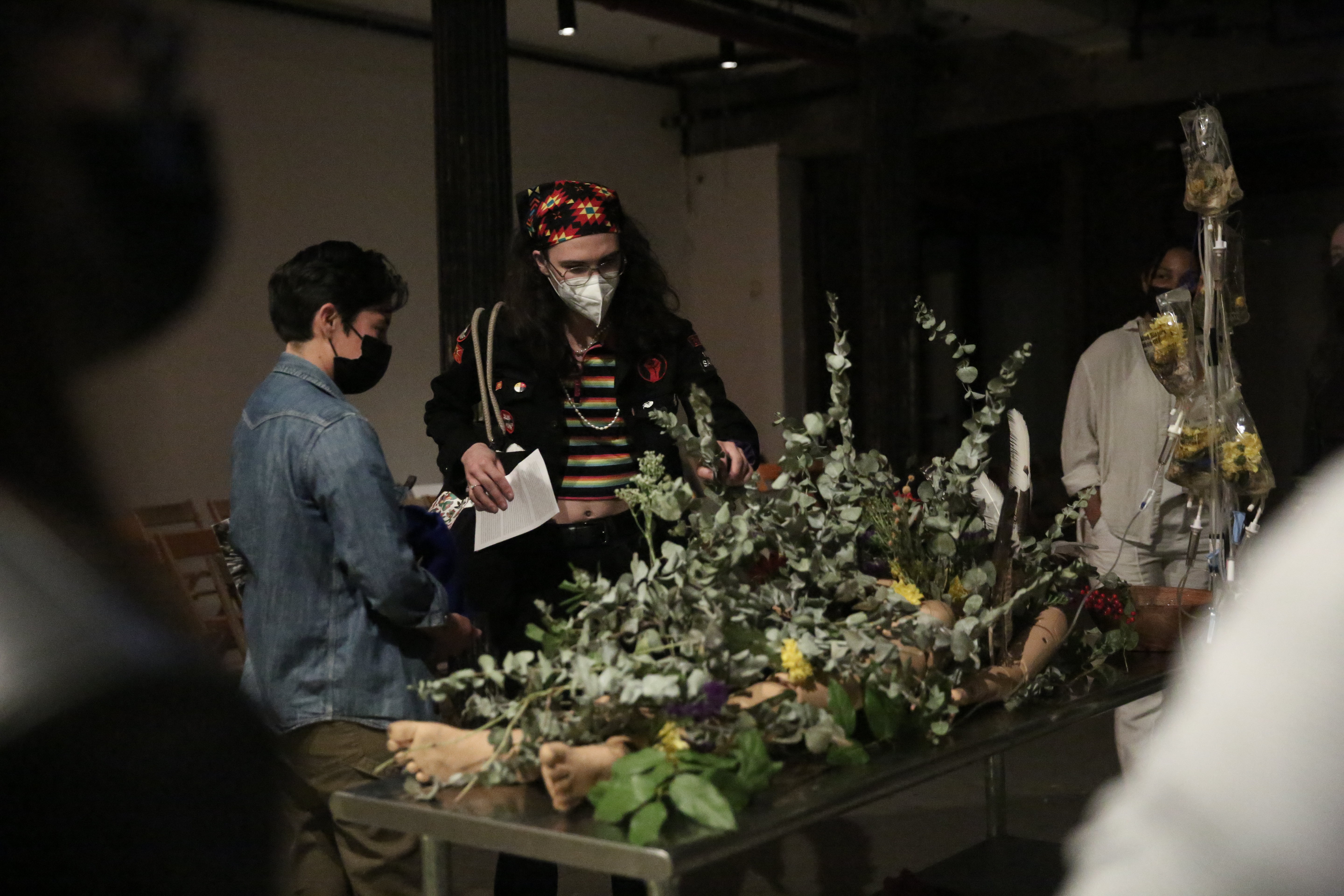 Various figures stand around a mannequin covered with foliage and flowers.