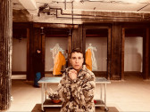 The artist devynn emory in their grandmother's fleece jacket covered in polar bears, standing in front of a silver sterile table. In the background is collaborator Joseph and cinematographer Jorge discussing where the ancestor forms draped in yellow fabric should be placed.
