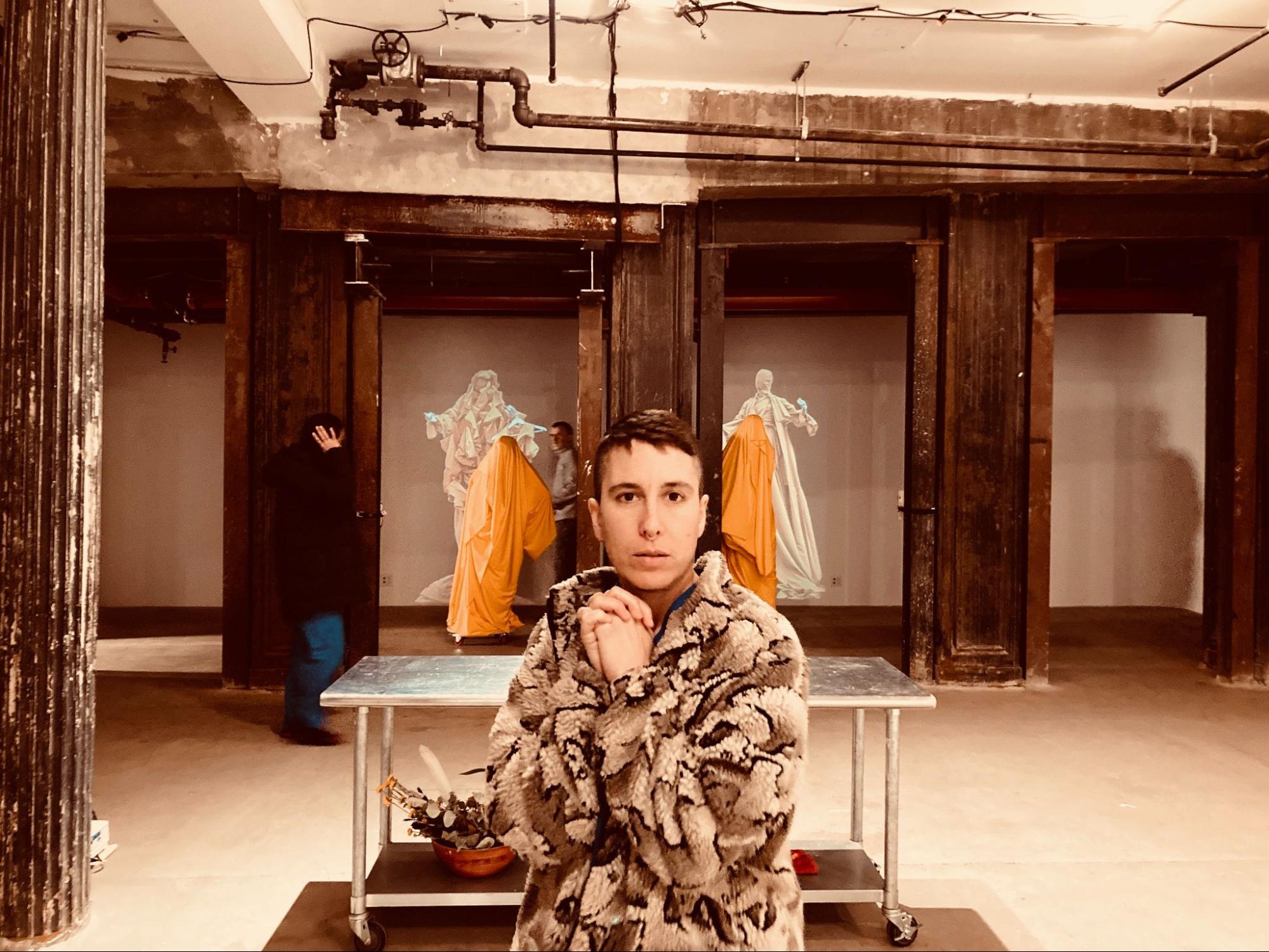The artist devynn emory in their grandmother's fleece jacket covered in polar bears, standing in front of a silver sterile table. In the background is collaborator Joseph and cinematographer Jorge discussing where the ancestor forms draped in yellow fabric should be placed.