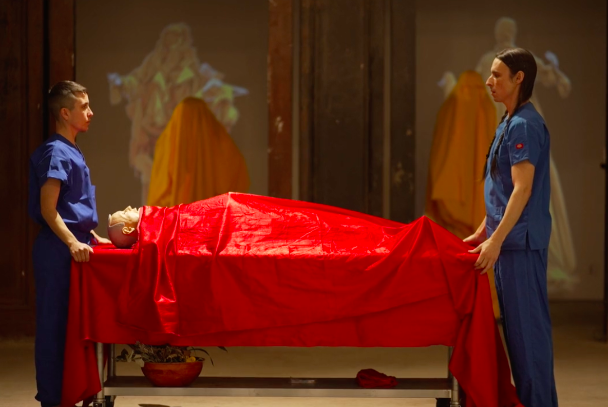 An image of two figures in blue scrubs standing on opposite ends of a sterile table. A medical mannequin on the table is covered in shiny red silk fabric. In the background, there are projections and forms draped in yellow fabric.