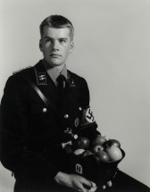 A black-and-white photograph of a man sitting down against a gray background wearing a Nazi uniform, holding a basket of apples.