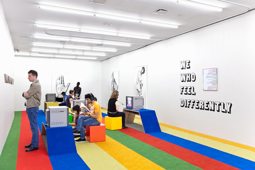 Installation photograph. The floor is covered with multicolored carpet strips, some of which are raised above the floor to create seating or shelving for CRT television monitors. On the wall, several black-and-white artworks are hung, depicting human figures, and text is printed on the wall 