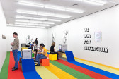 Installation photograph. The floor is covered with multicolored carpet strips, some of which are raised above the floor to create seating or shelving for CRT television monitors. On the wall, several black-and-white artworks are hung, depicting human figures, and text is printed on the wall 