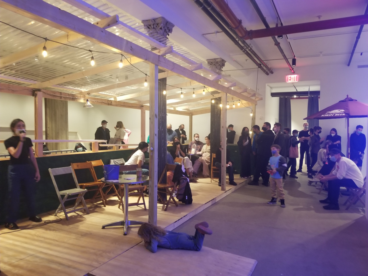 A green and tan wooden dining corral with a ramp, metal tables and chairs. People sit and stand in and around the corral.