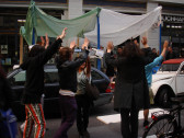 A group of people dressed in black standing in the street with their hands up. Behind them, green and white fabrics on top of blue poles create a tent-like structure. The word 