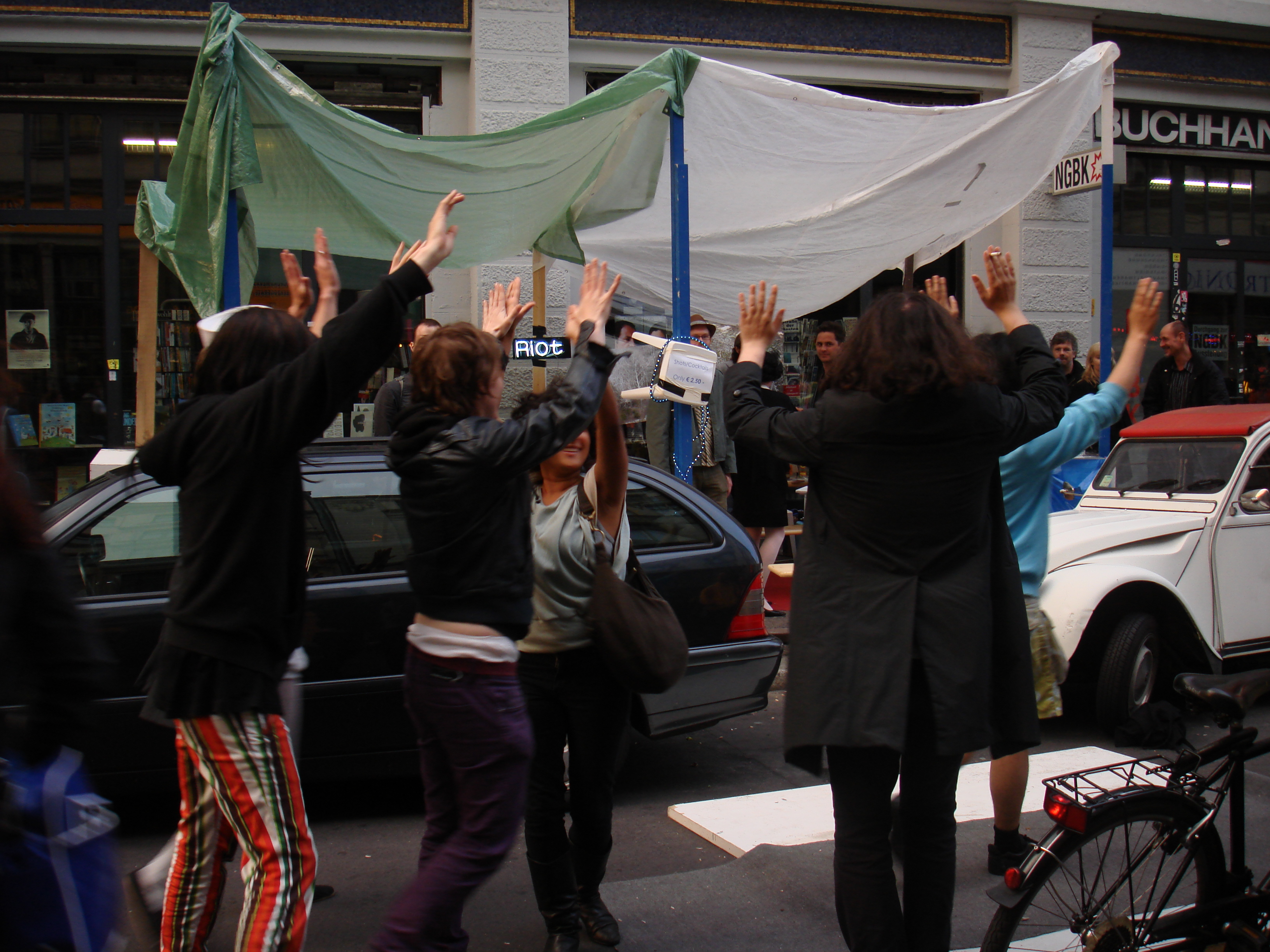 A group of people dressed in black standing in the street with their hands up. Behind them, green and white fabrics on top of blue poles create a tent-like structure. The word 