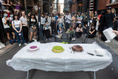 Three cakes sit atop a white table in an alleyway. A crowd behind a black net looks on intently.