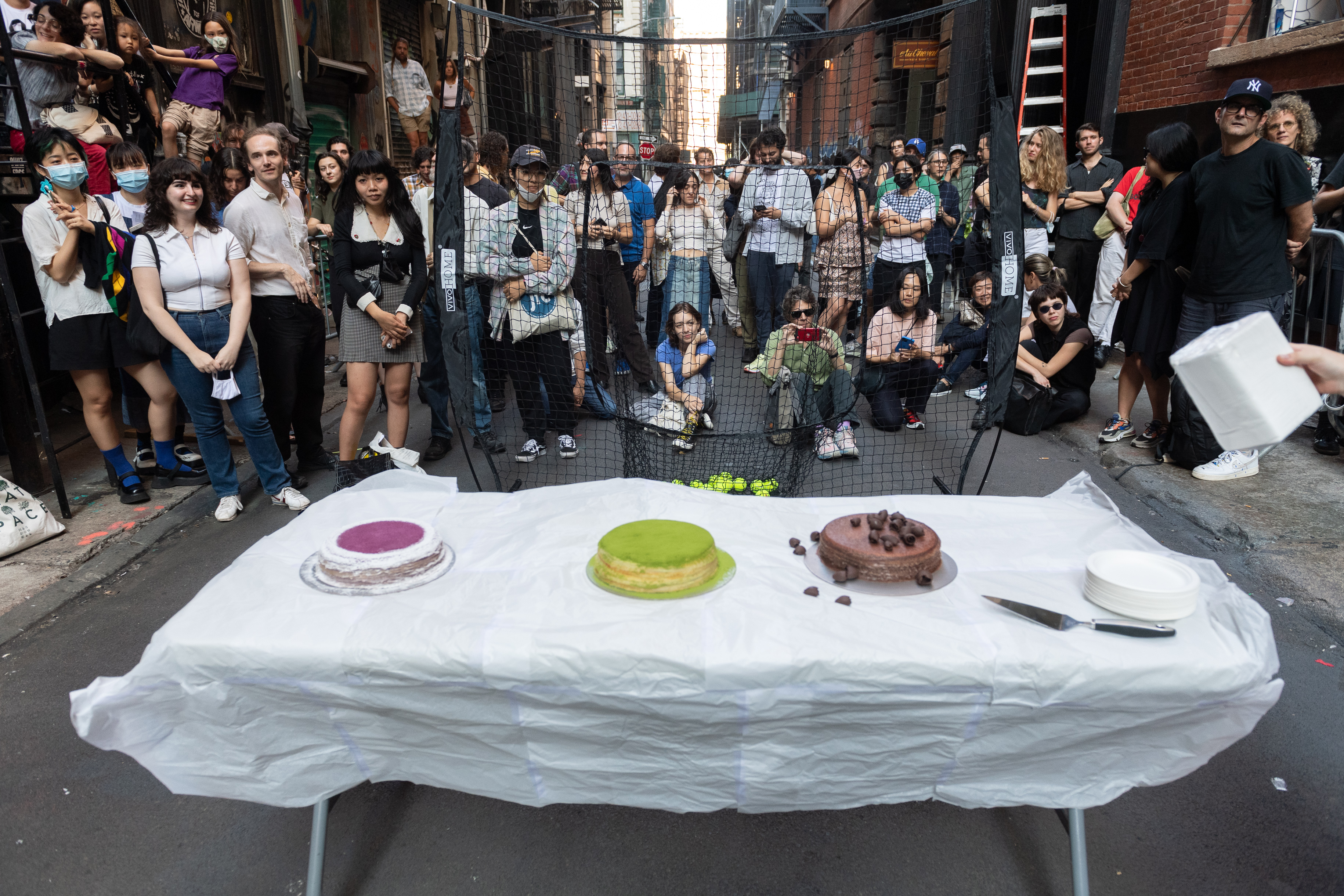 Three cakes sit atop a white table in an alleyway. A crowd behind a black net looks on intently.