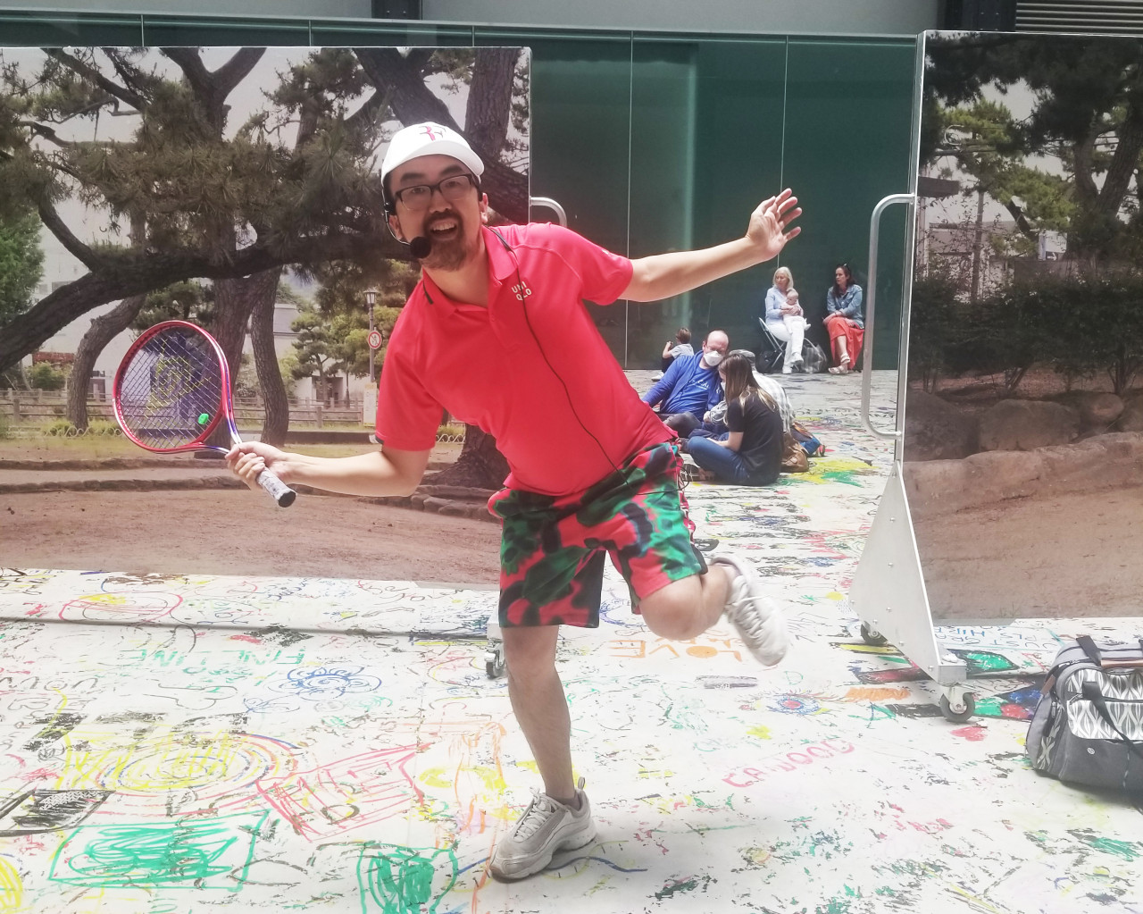 A person in a red shirt, white cap, and multicolored pants smiles and poses with a tennis racquet, standing atop a large piece of paper with colorful drawings on it. In the background are people sitting on the ground and printed images of trees.