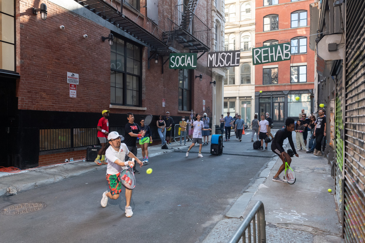 Six people with tennis rackets hit tennis balls against a graffitied metal door in an alleyway. Suspended behind them are a black net and signs that read "Social Muscle Rehab" in painted white