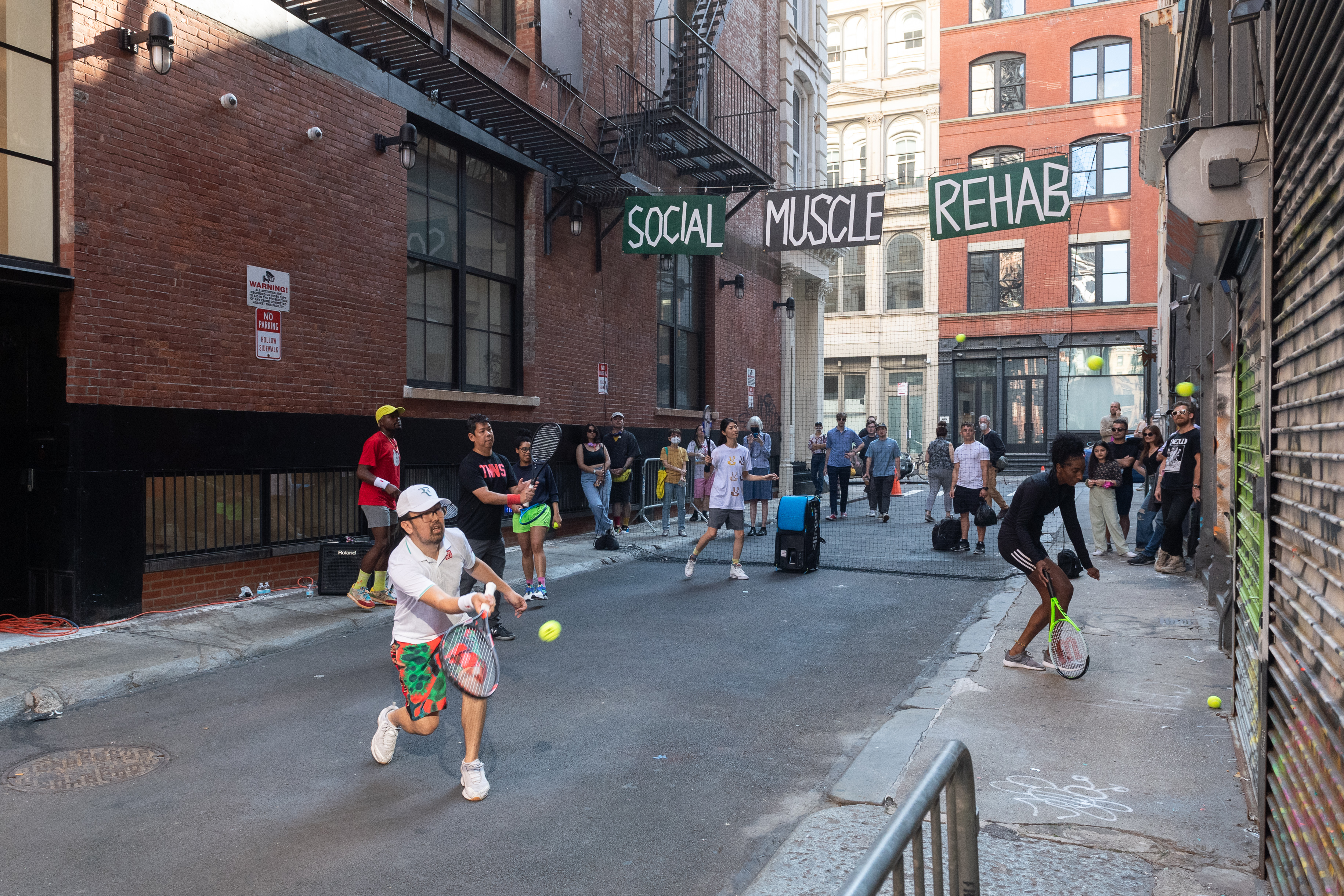Six people with tennis rackets hit tennis balls against a graffitied metal door in an alleyway. Suspended behind them are a black net and signs that read 