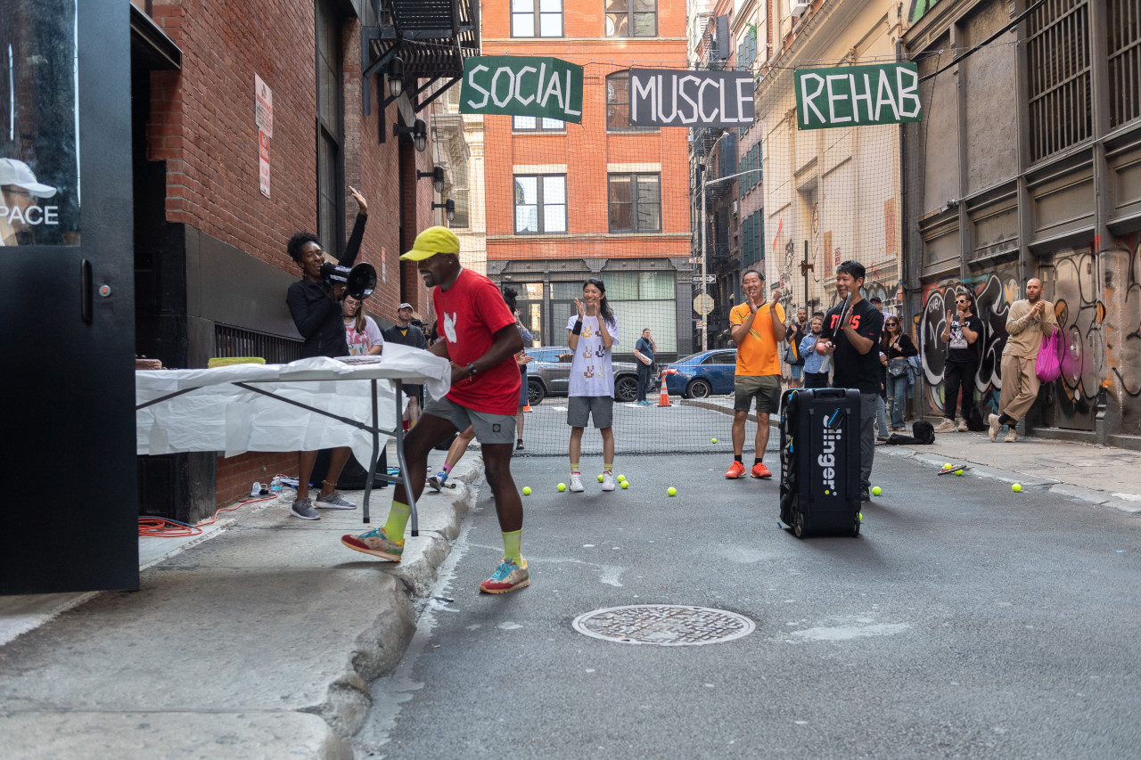 Two people carry a white table out of a door and into an alleyway. Behind them several others cheer, one with a megaphone. Suspended in the background are a black net and black and green signs that read "Social Muscle Rehab" in white painted text.