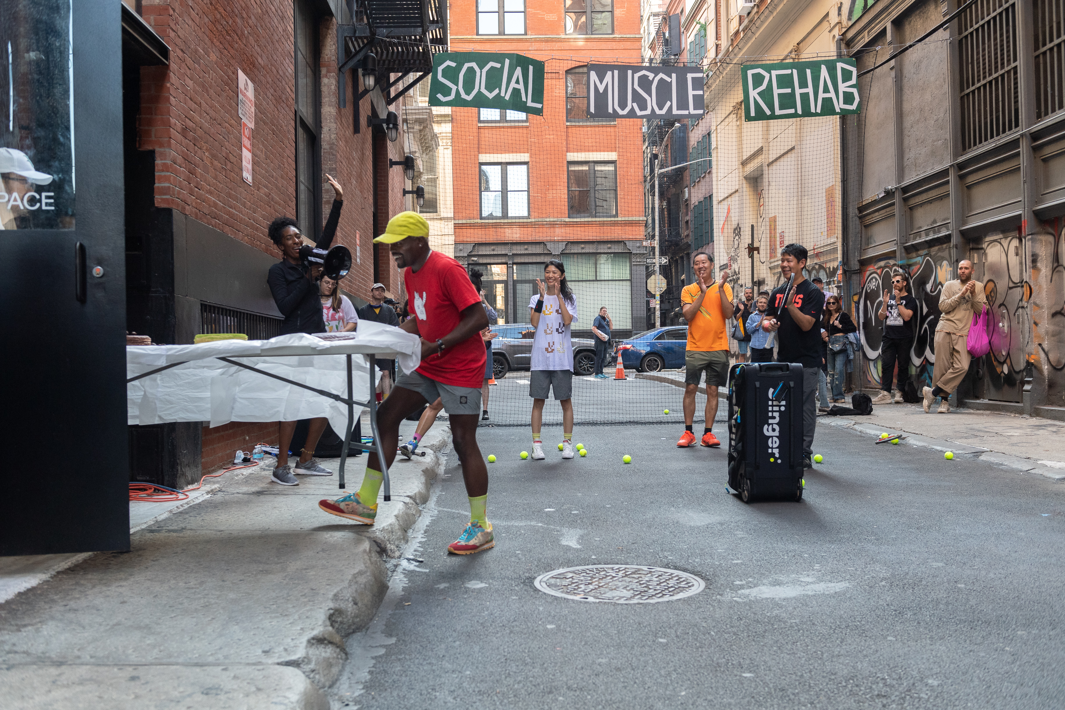 Two people carry a white table out of a door and into an alleyway. Behind them several others cheer, one with a megaphone. Suspended in the background are a black net and black and green signs that read 