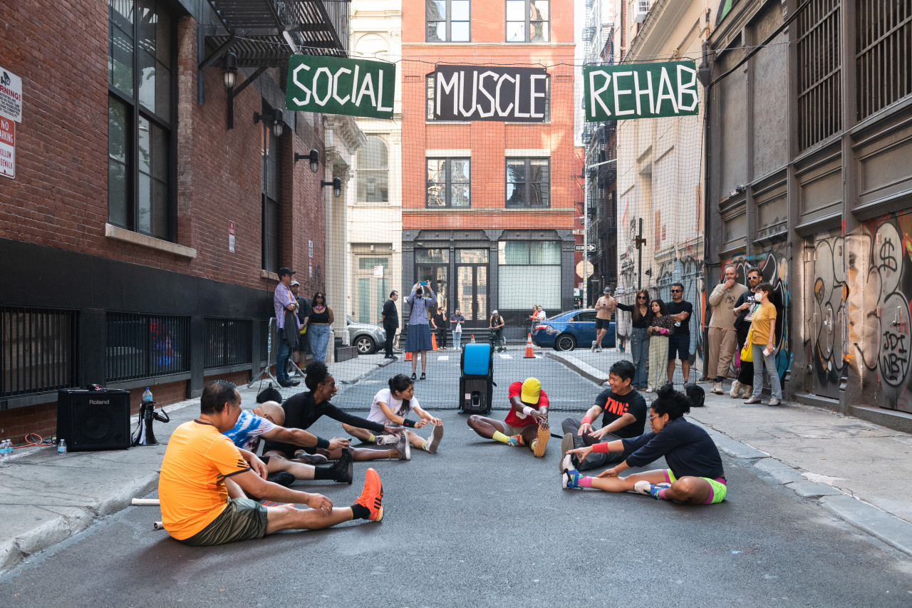 Six people stretch on the ground of an alleyway. Suspended behind them, the words "Social Muscle Rehab" are painted in white on black and green signs.