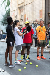 Four people stand in an alleyway in front of a black net, one of them holding a black megaphone. Five tennis balls lie at their feet.