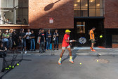 A person with a yellow hat and red shirt hits a tennis ball in an alleyway. A small crowd watches in the background on the left.