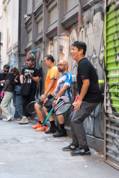 Three people holding tennis rackets squat against a grey graffitied metal door in an alleyway, pressing their backs against green tennis balls.