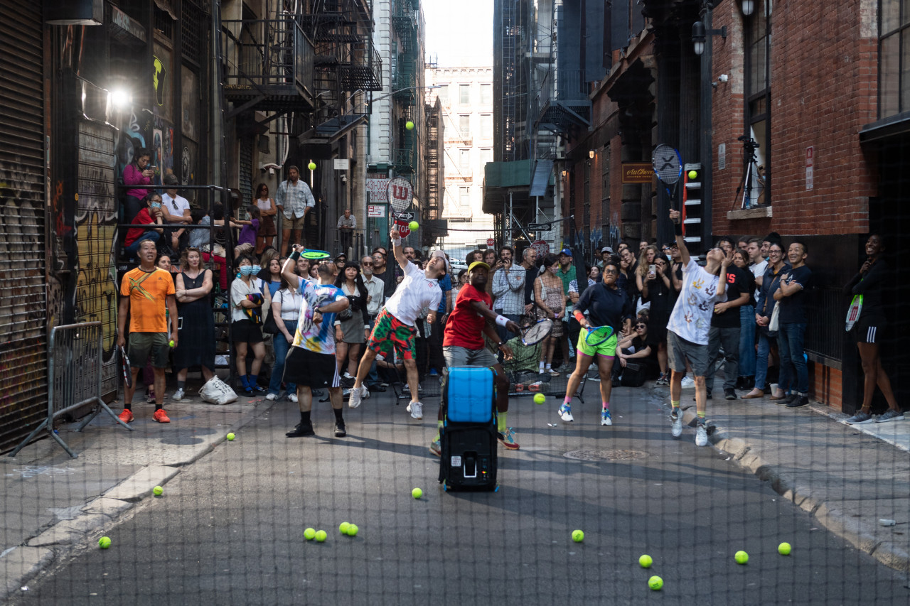 Six people with tennis rackets in an alleyway jump and swing to hit tennis balls towards the camera which is behind a black net. A crowd behind the tennis players watches attentively.
