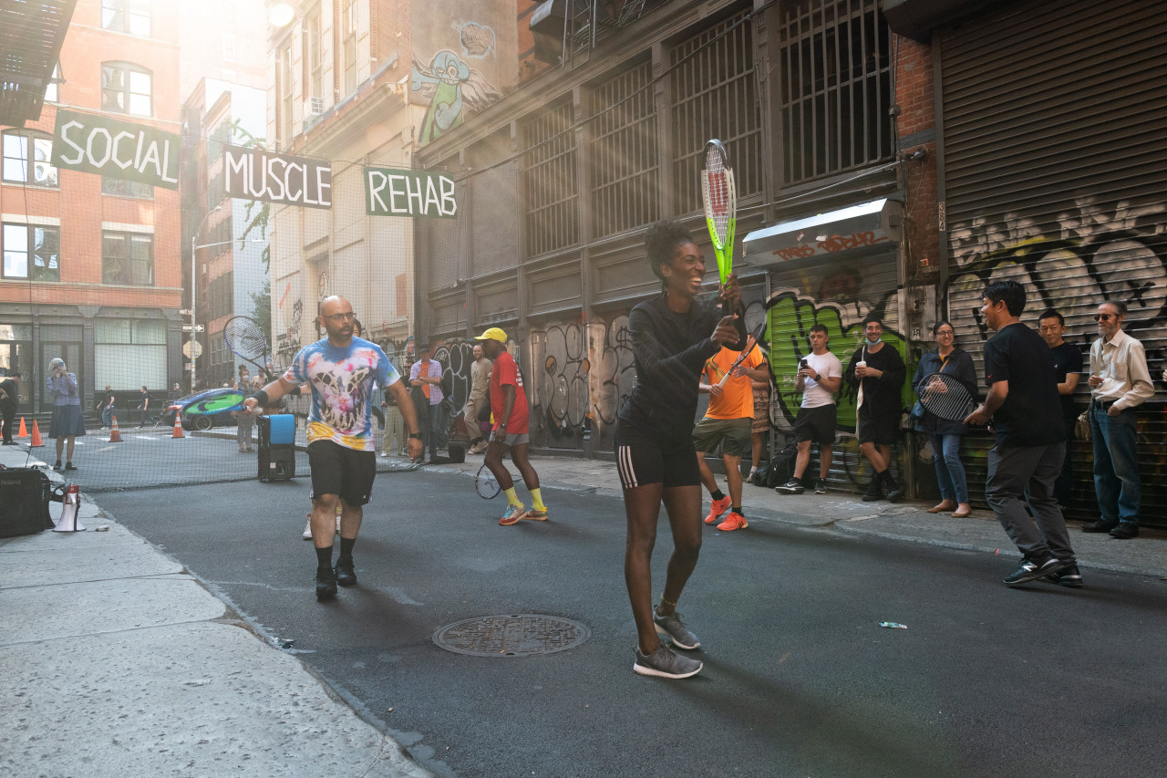 Five people holding tennis rackets running in a circle in an alleyway. A small crowd watches, standing against a brick wall with metal doors covered in graffiti on the right.