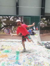 A person in a red shirt, white cap, and multicolored pants smiles and poses with a tennis racquet, standing atop a large piece of paper with colorful drawings on it. In the background are people sitting on the ground and printed images of trees.