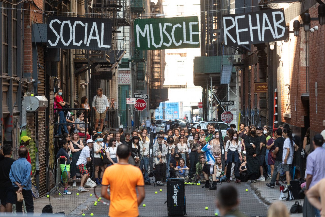 Tennis players stand in an alleyway, ready to hit balls from an automatic ballfeeder. A crowd watches intently in the background. Suspended above them the words "Social Muscle Rehab" are painted in white capitalized text on black and green signs.
