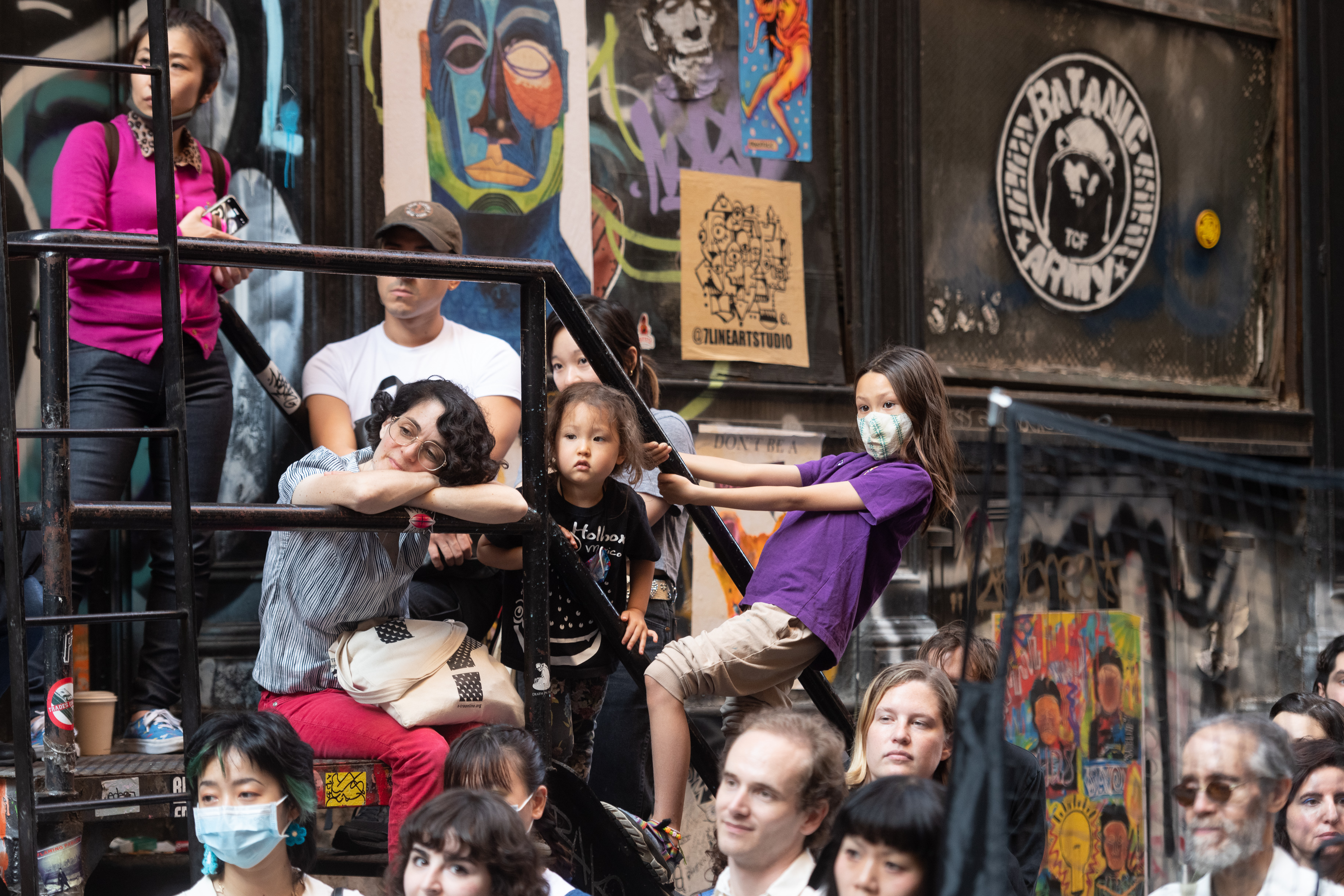 A crowd looking left in an alleyway, a child in a purple shirt and mask holds onto a balcony railing leaning out over the crowd to get a better view.
