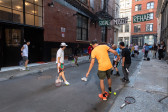 Six people holding tennis rackets stand in an alleyway. Each of them has their right shoe off and rubs the sole of their foot on top of a tennis ball. Suspended in the background, the words 