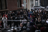 A crowd of people standing outside in Cortlandt Alley surrounding several performers. The performers are mixed in with the crowd, holding metal poles, some looking at a baby doll which is photographed in midair after being thrown upwards.