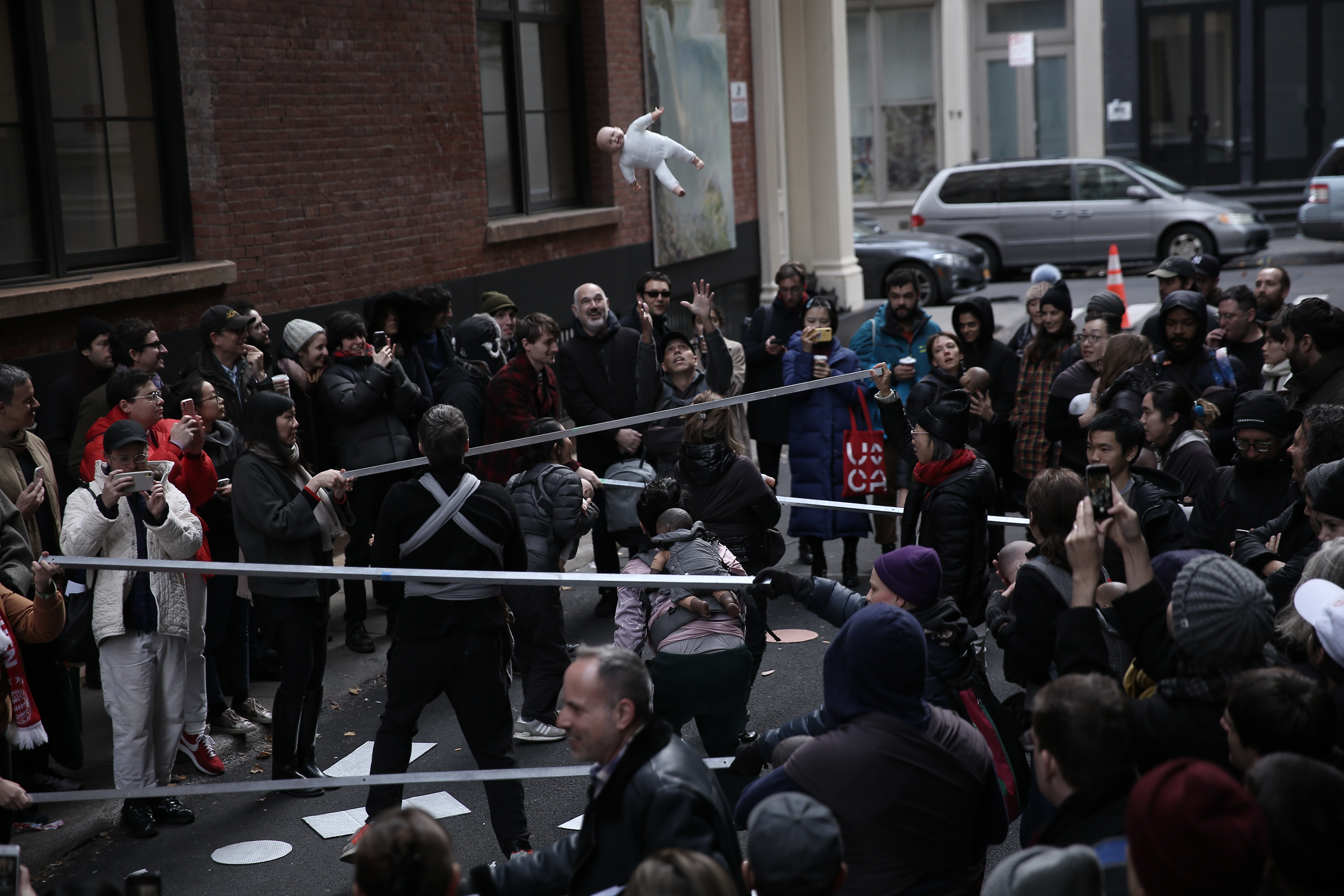 A crowd of people standing outside in Cortlandt Alley surrounding several performers. The performers are mixed in with the crowd, holding metal poles, some looking at a baby doll which is photographed in midair after being thrown upwards.