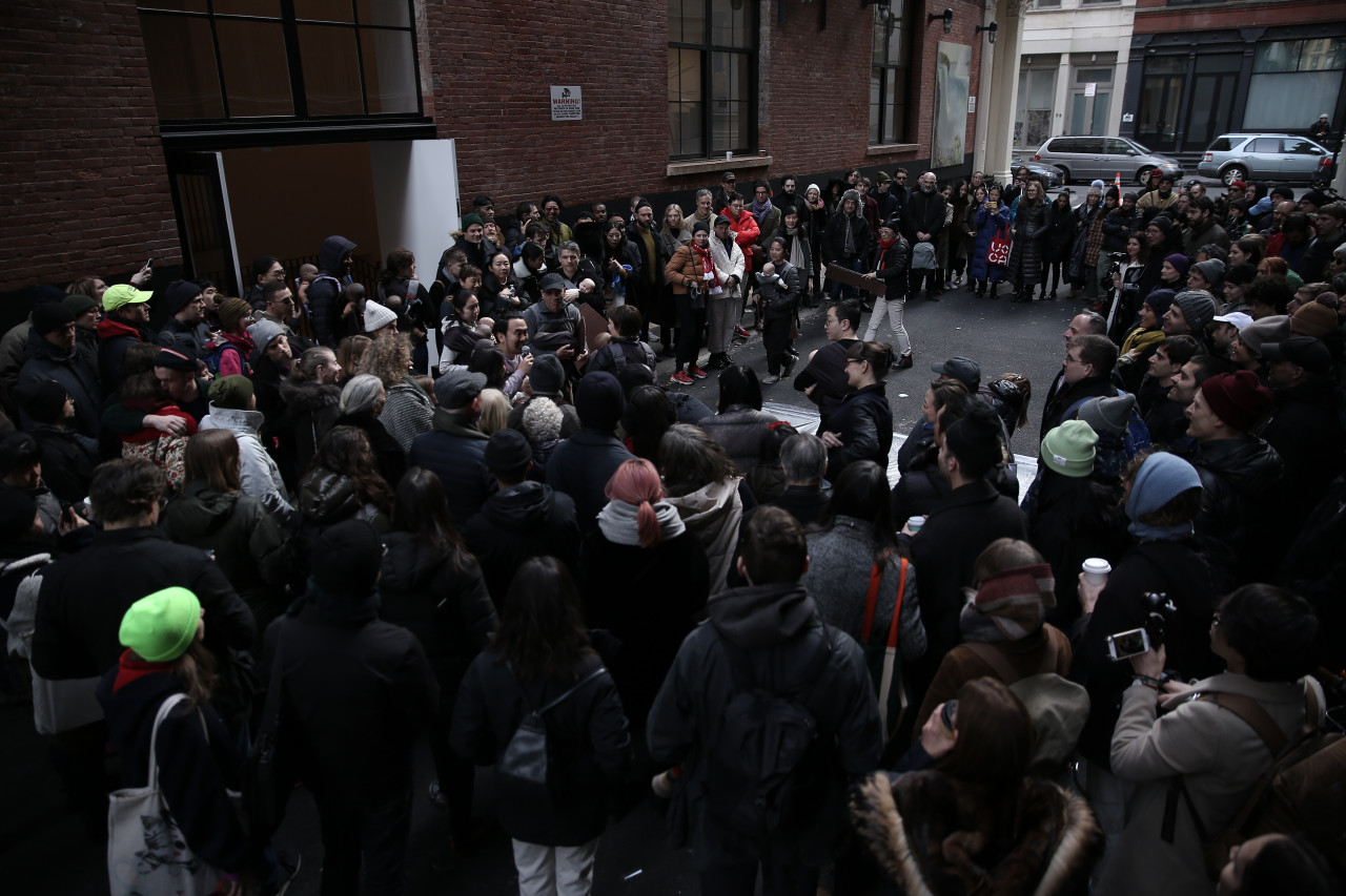 A large group of people photographed from above, stading in Cortlandt Alley, forming a small circle in the middle. Some people standing near the center hold baby dolls.