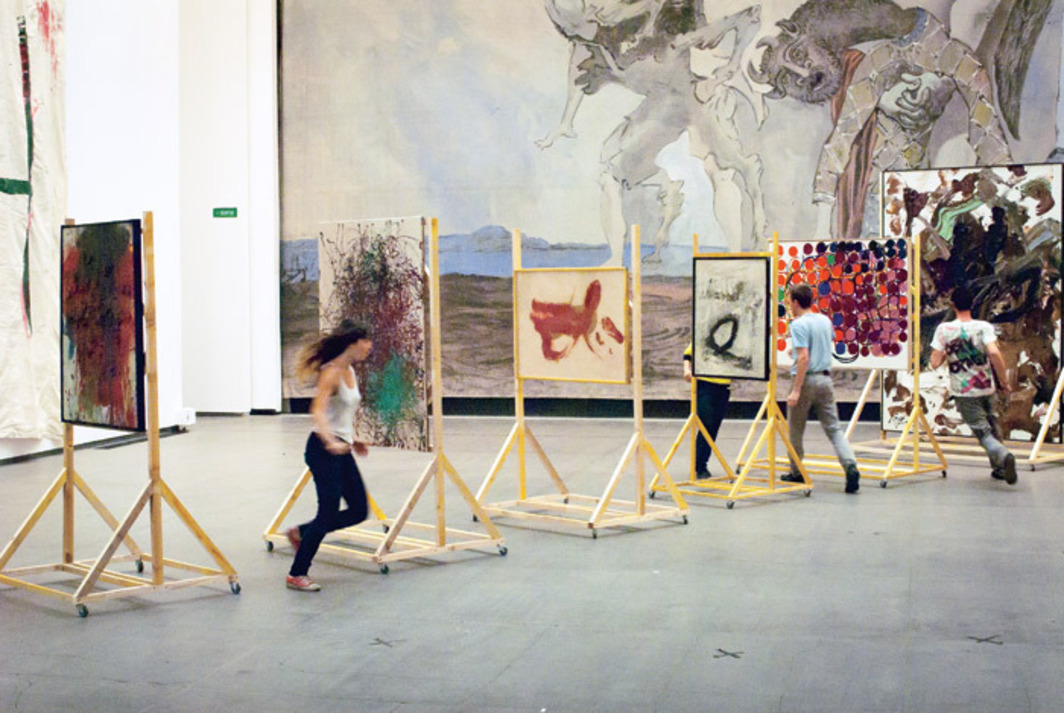 An image of several people running between six large, abstract paintings on wooden stands arranged diagonally through a room. A large painting covers the back wall, and a large sheet of fabric hangs loosely from the left wall, with a minimal amount of green and red paint on it.