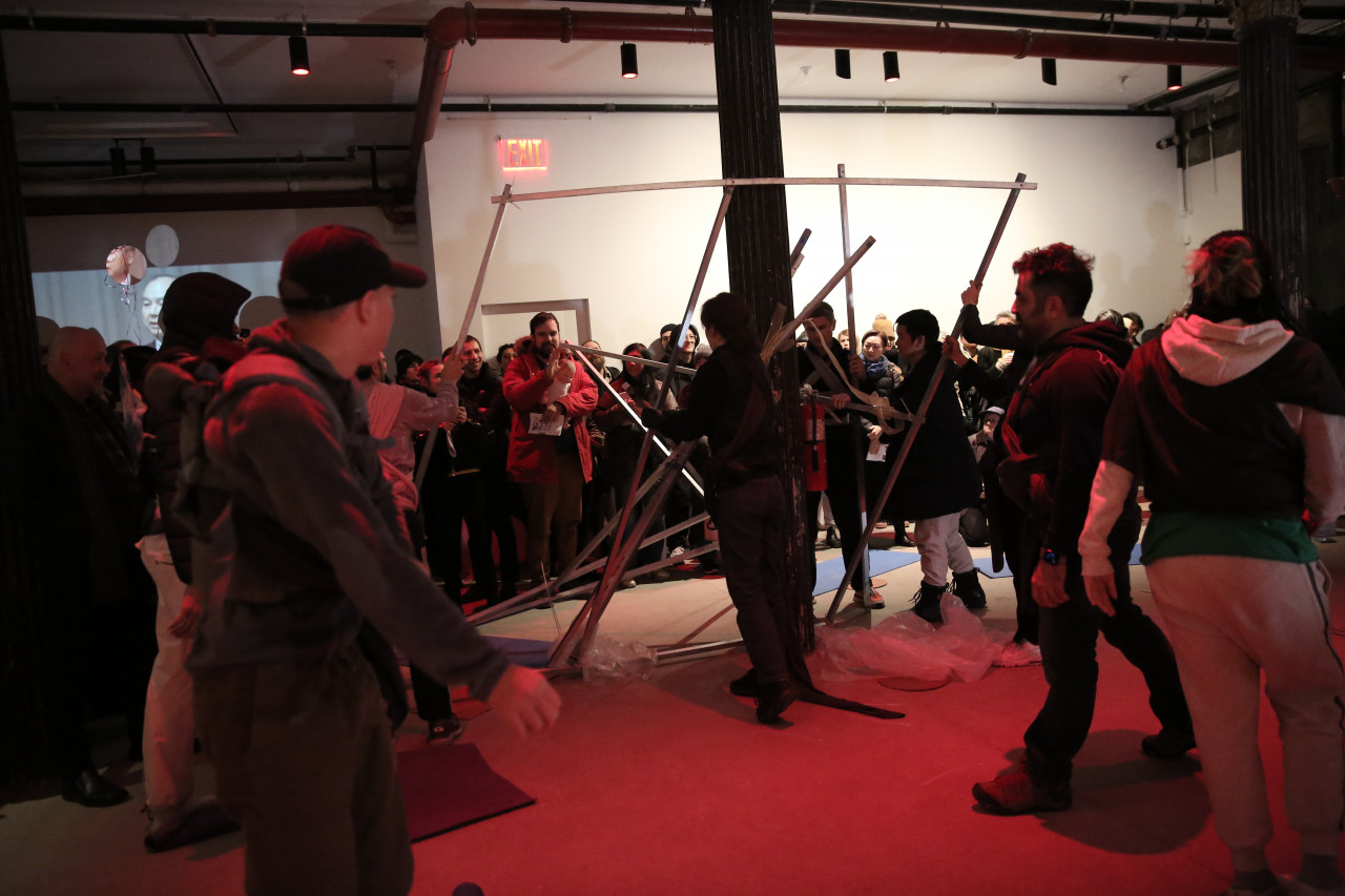 Several people hold a makeshift metal structure upright, fastening additional metal poles onto the structure. A small crowd stands behind them watching.