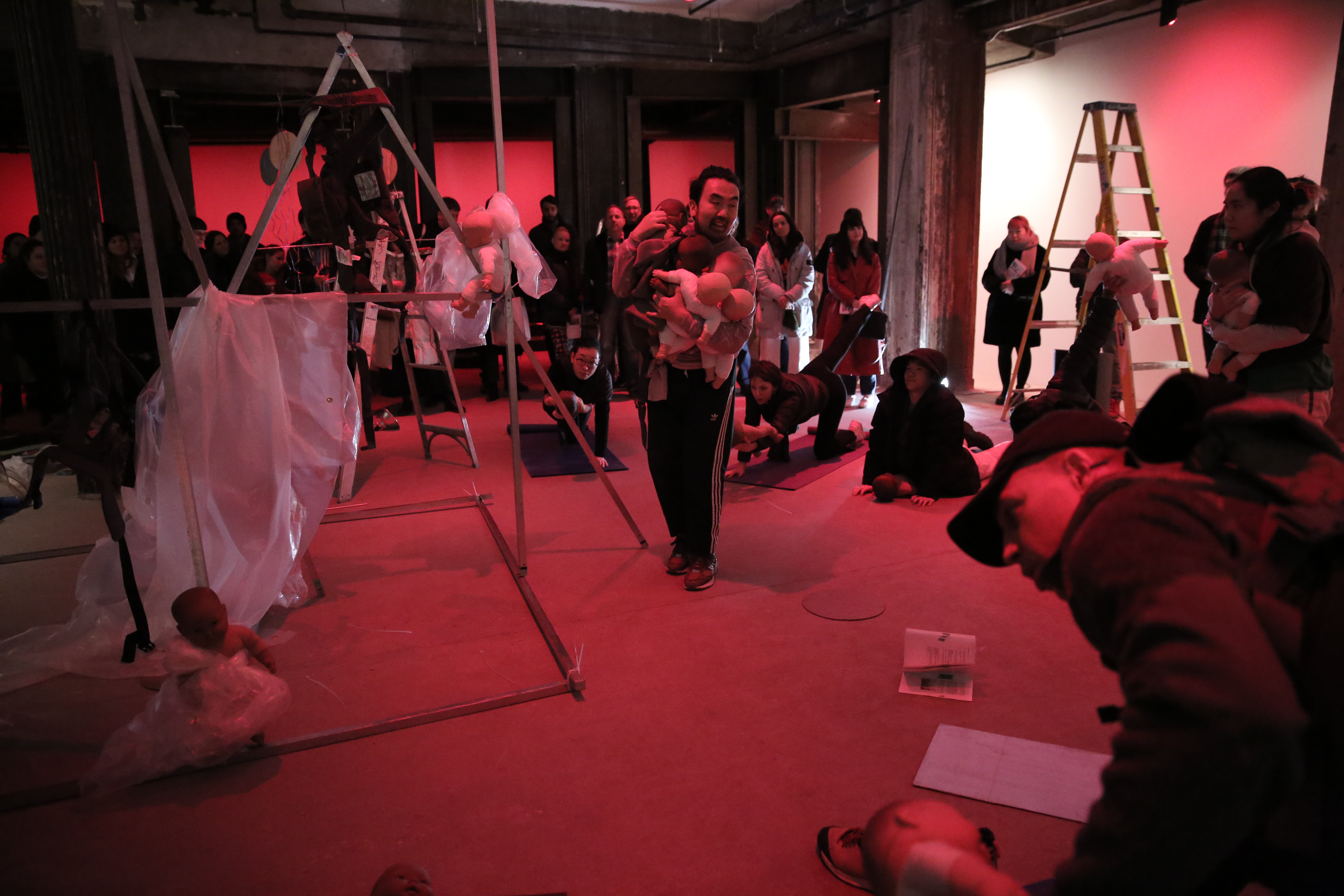 A man stands in the center of a room holding numerous baby dolls, surrounded by several performers striking yoga poses and a small crowd. A makeshift geometric metal structure stands next to him, with a baby doll sitting in a crease of the structure.