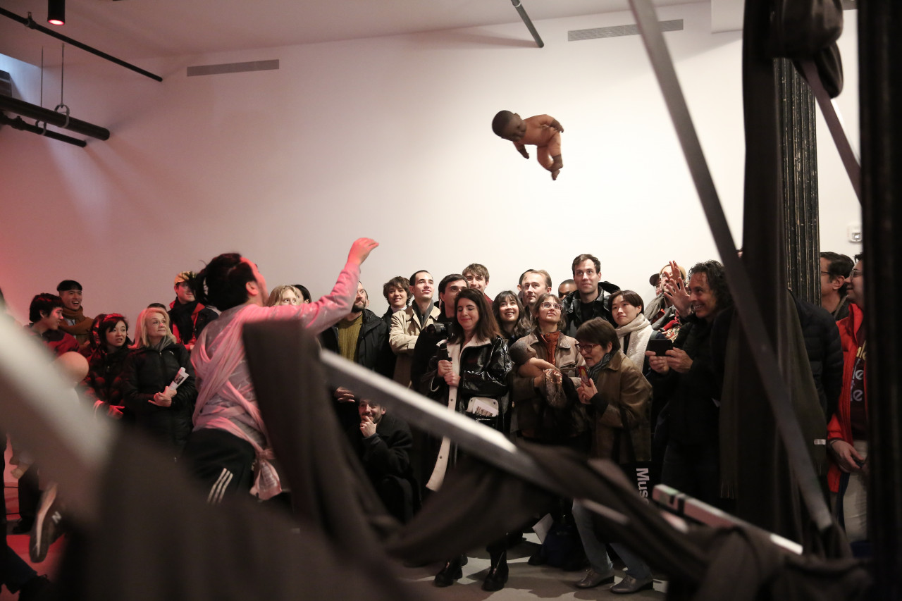 A man gazes up at a baby doll mid-flight, holding out his right hand towards it. A group of people gaze towards the doll, smiling.