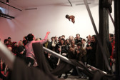A man gazes up at a baby doll mid-flight, holding out his right hand towards it. A group of people gaze towards the doll, smiling.