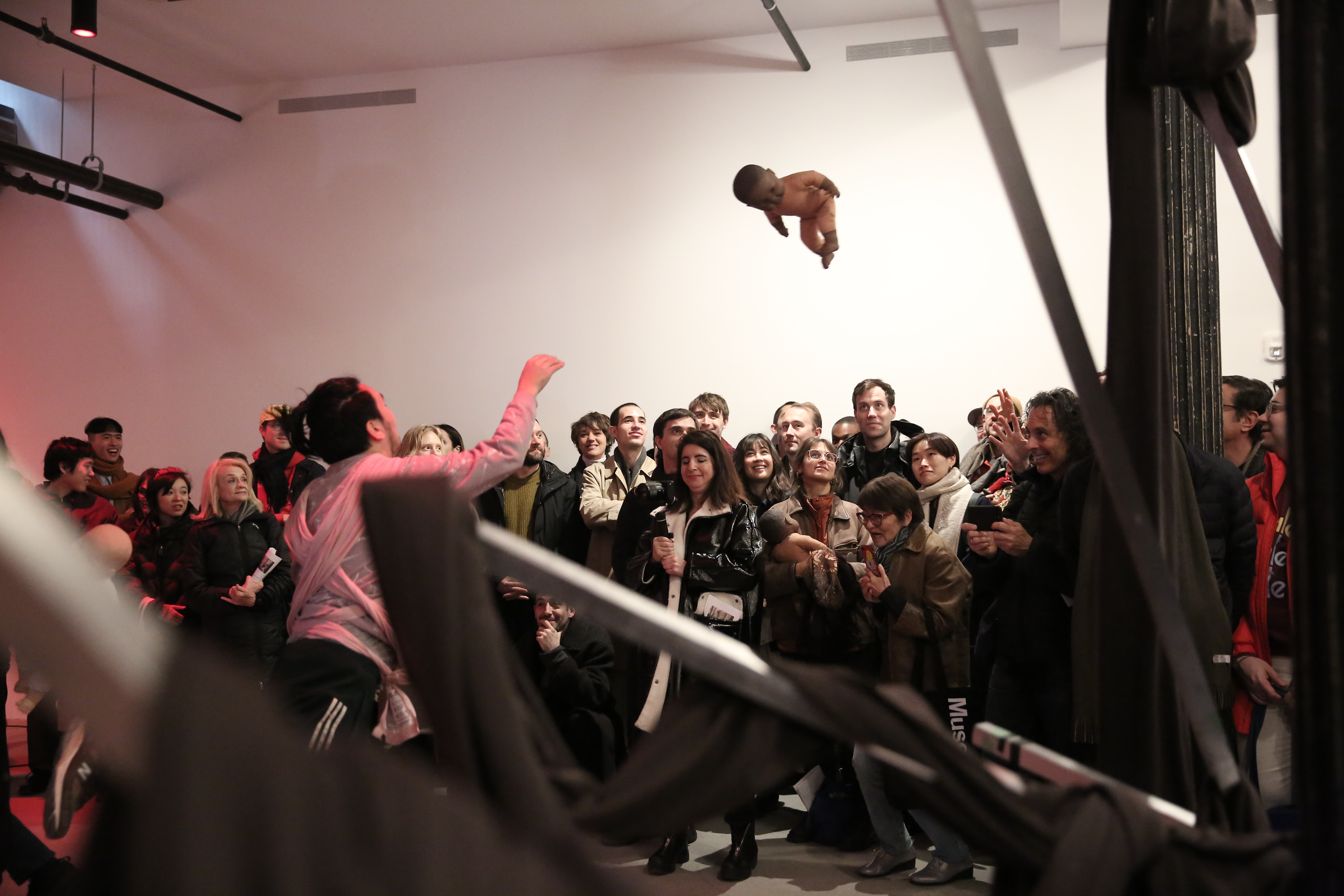 A man gazes up at a baby doll mid-flight, holding out his right hand towards it. A group of people gaze towards the doll, smiling.