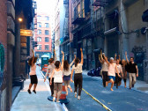 A photo of a city alleyway acts as the background for this image. Superimposed on the photo are two groups of people in motion with right arms raised, holding cellphones. The group on the right faces toward the viewer, the group on the left faces away.
