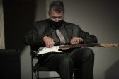 A seated figure plays a guitar that lies in his lap.