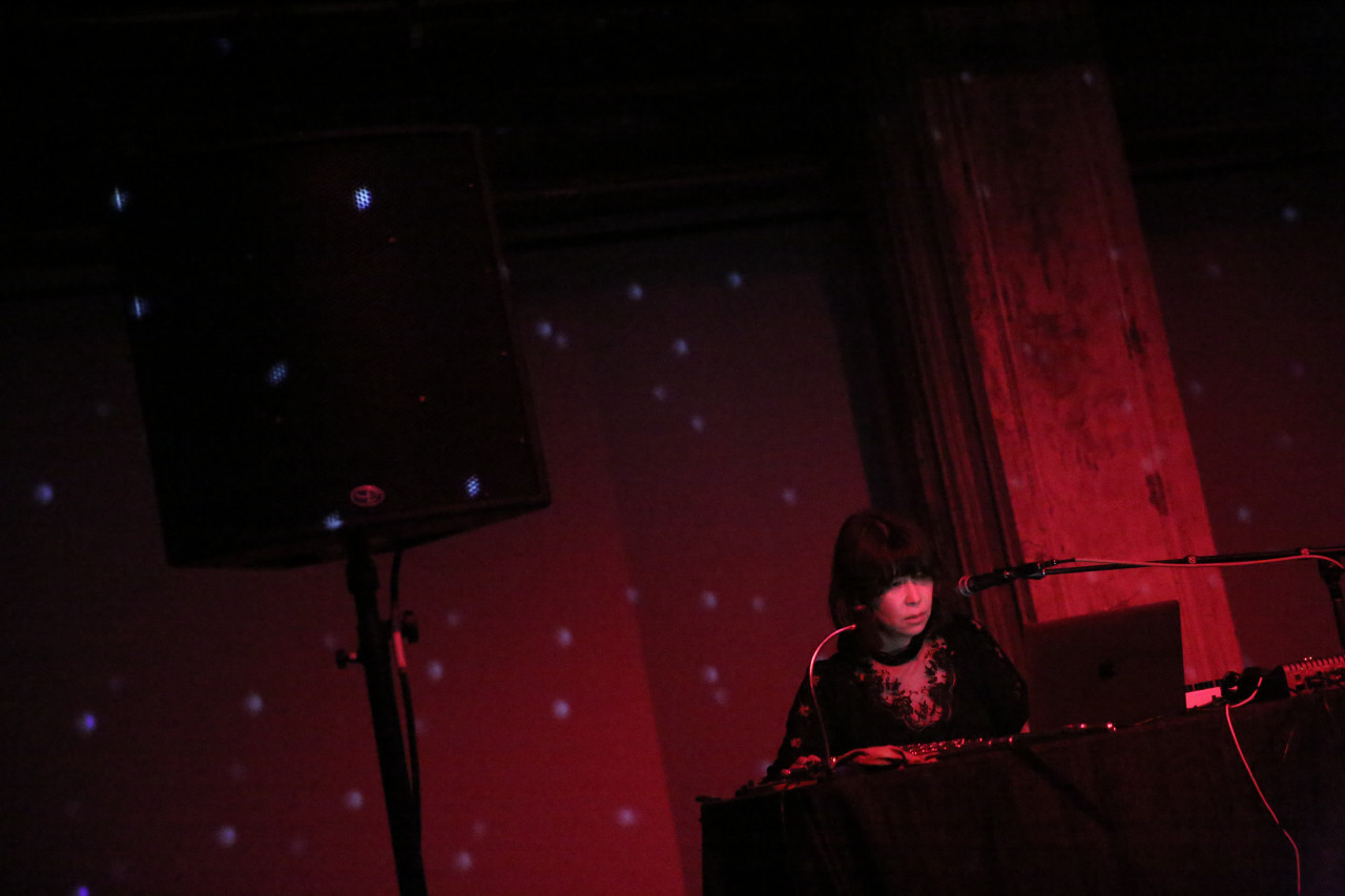A figure peers into a computer while standing behind a table and next to a speaker.