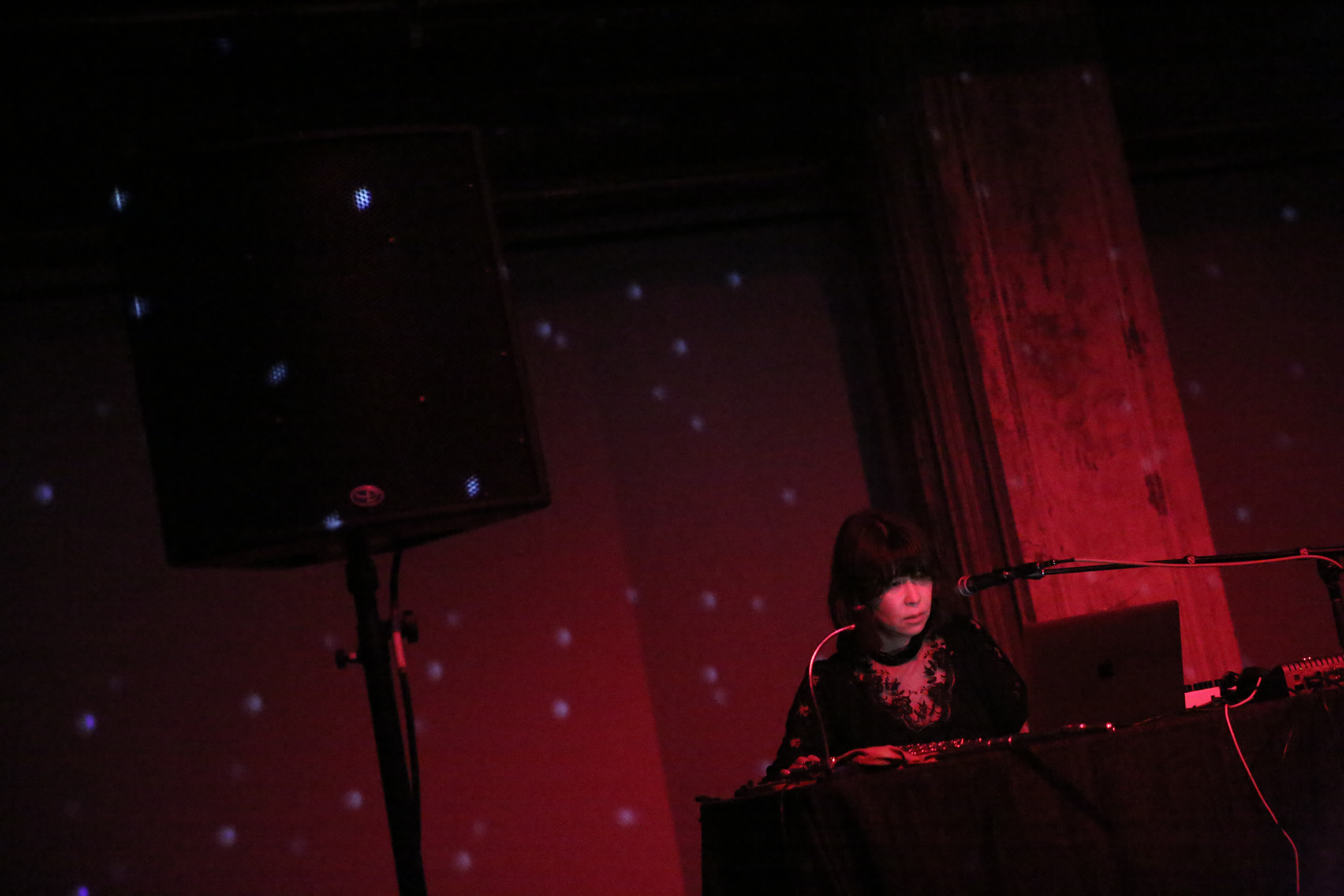 A figure peers into a computer while standing behind a table and next to a speaker.