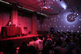 A seated crowd watches a performer who stands behind a table on a stage. The room is illuminated by red light and the reflection of a disco ball.