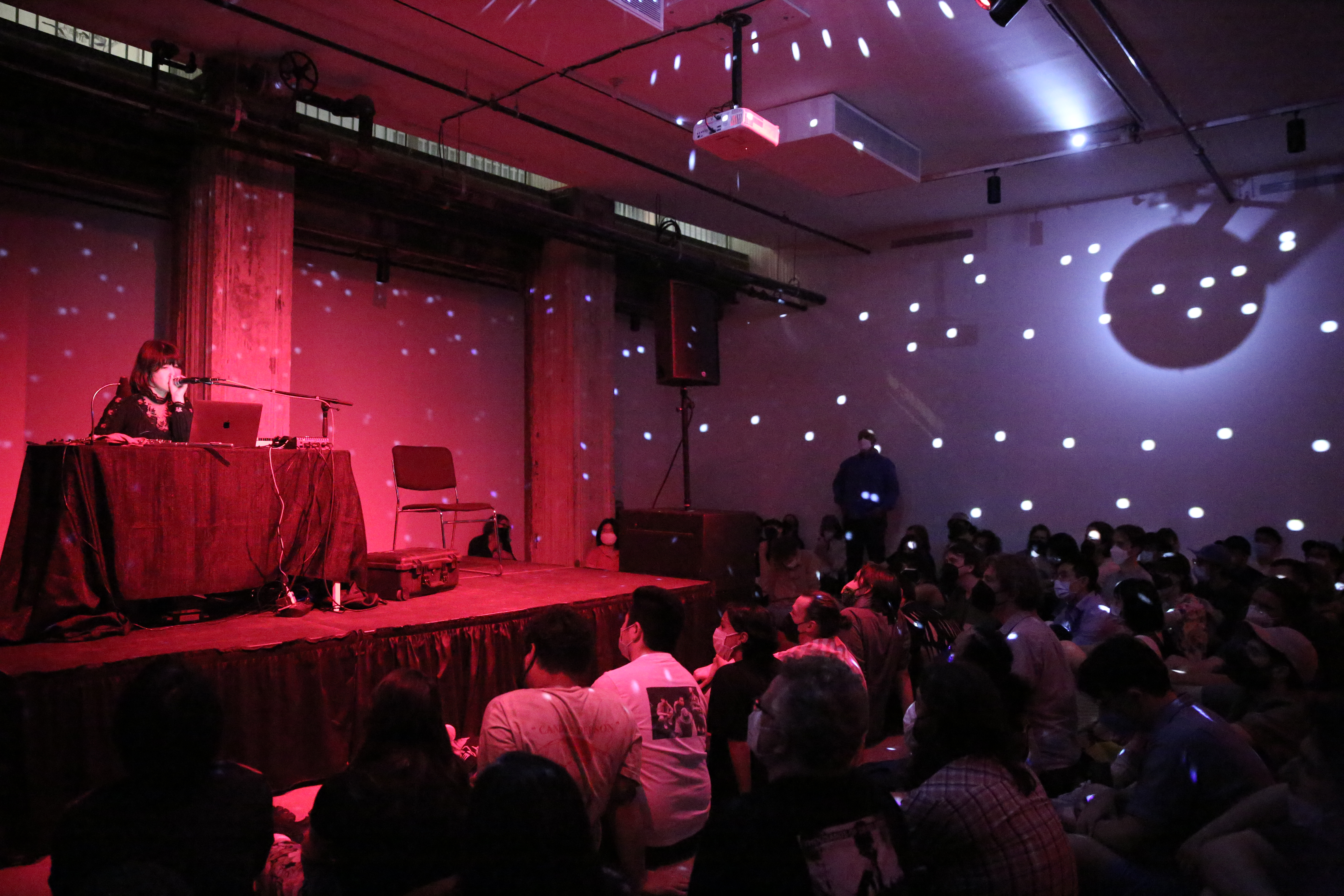 A seated crowd watches a performer who stands behind a table on a stage. The room is illuminated by red light and the reflection of a disco ball.