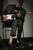 A figure wearing a suit plays guitar, their feet dangling over a pedal.