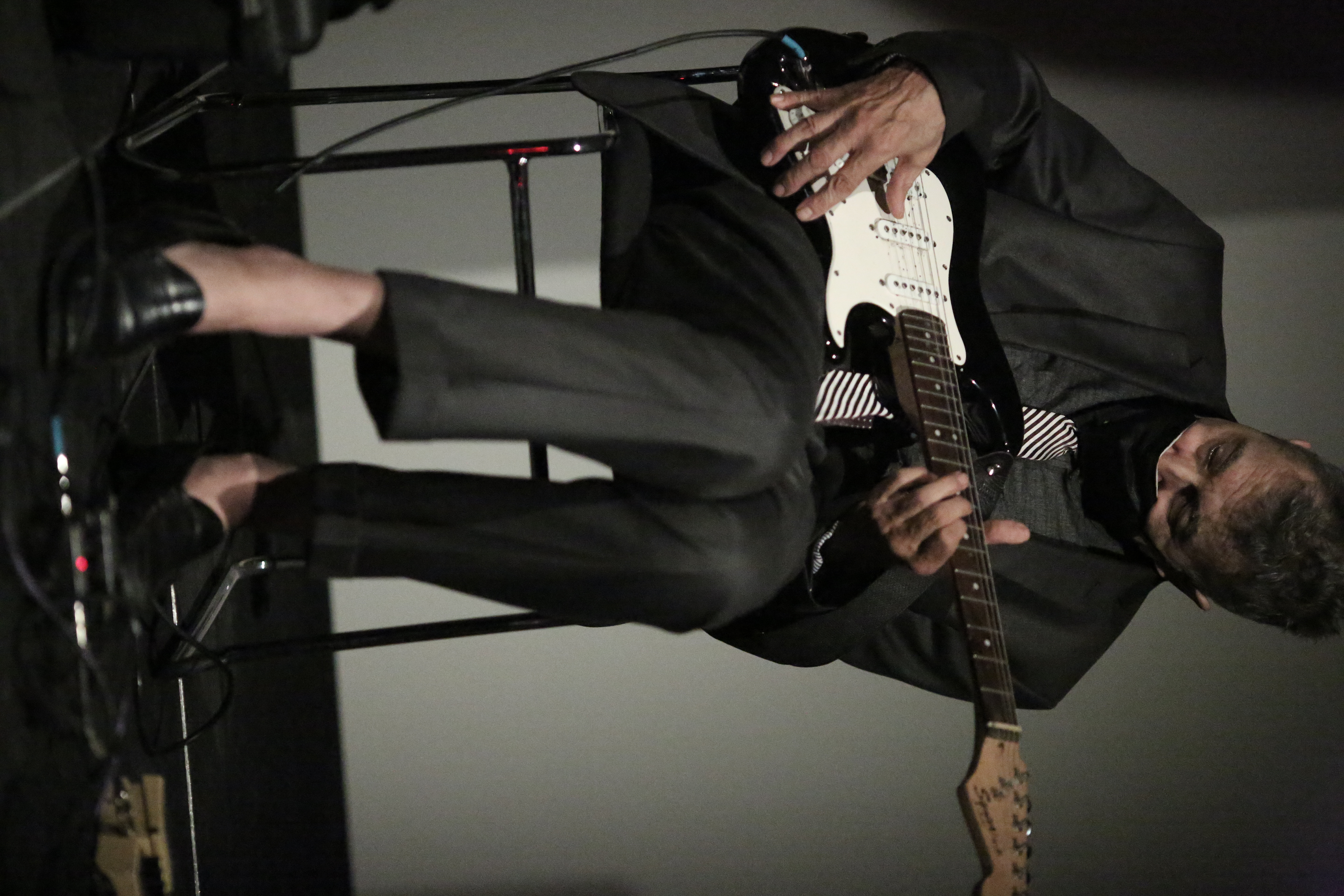 A figure wearing a suit plays guitar, their feet dangling over a pedal.