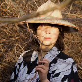 A color photograph of a figure in a pointed hat with a wide brim and wearing a black and white robe. The figure is pictured amid tree branches and vines. They hold a twisted vine in their left hand.
