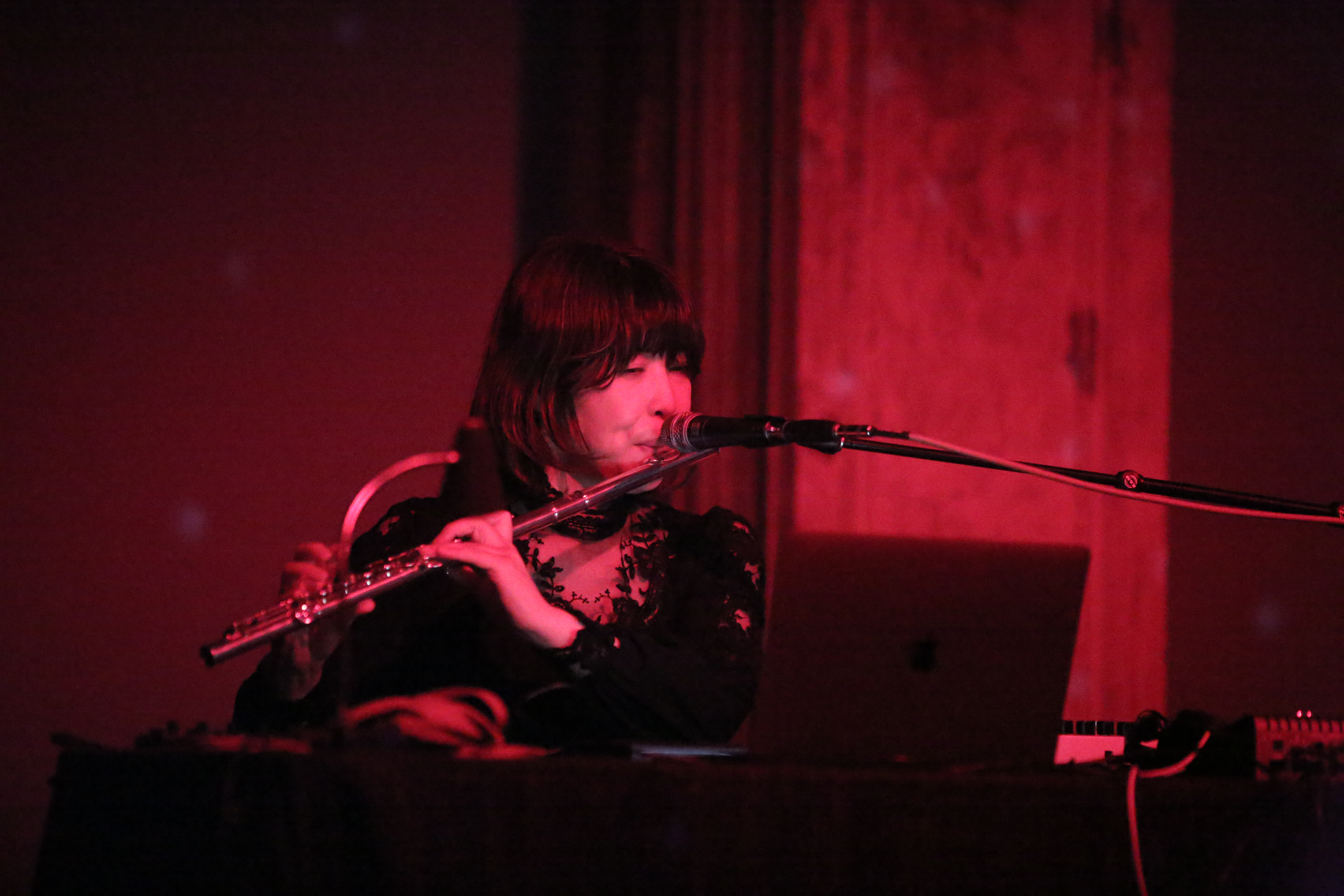A figure whose face is illuminated by red light plays flute into a microphone.