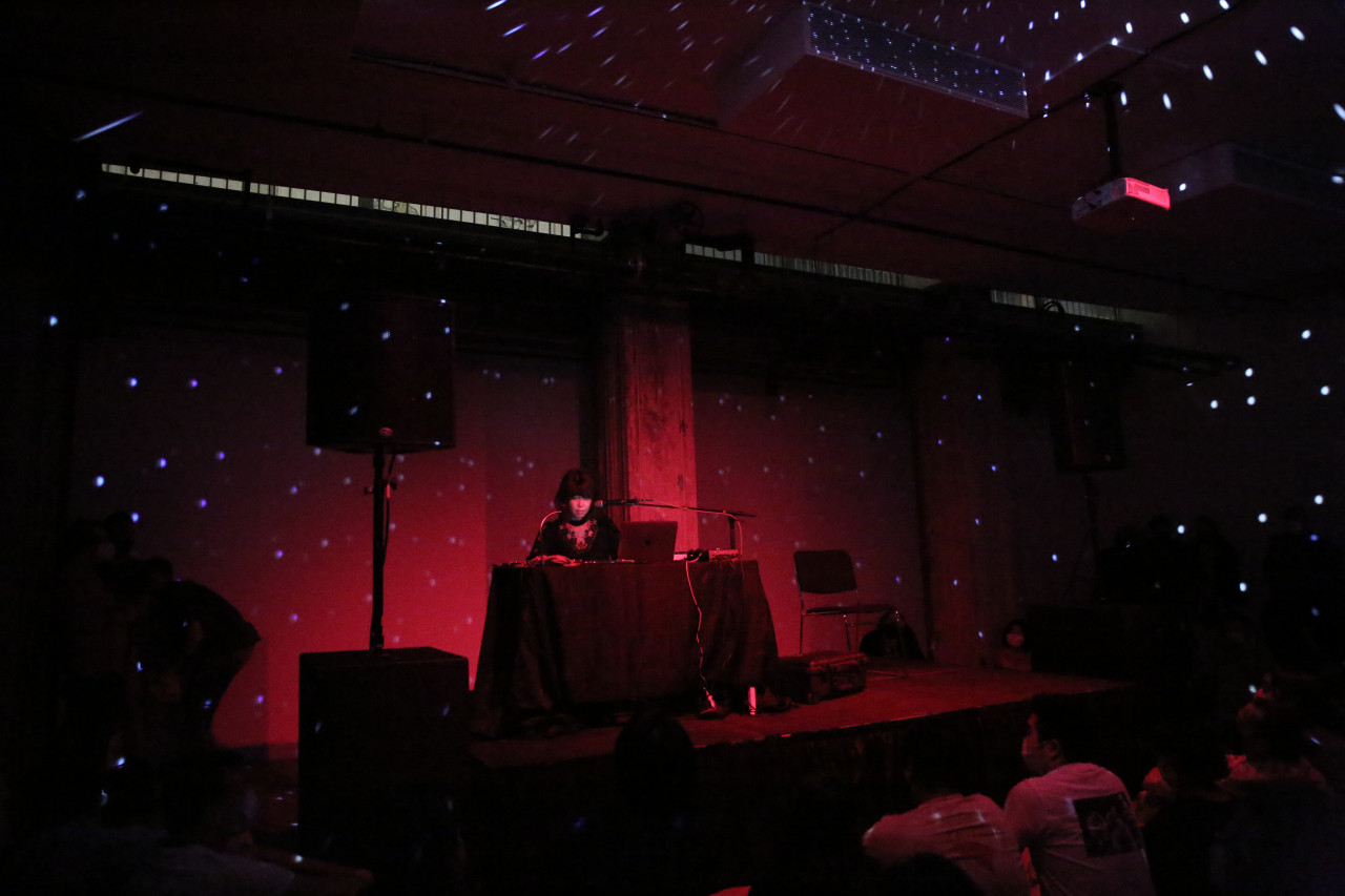 A figure stands behind a table on a stage. Across the room, red light and small fragmented pieces of white light can be seen.