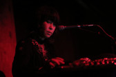 A woman stands behind a microphone under dim red light. The woman looks down to her right while typing on a keyboard.